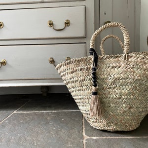 May include: A large, woven straw tote bag with black and white tassel accents. The bag is sitting on a tiled floor in front of a white dresser.