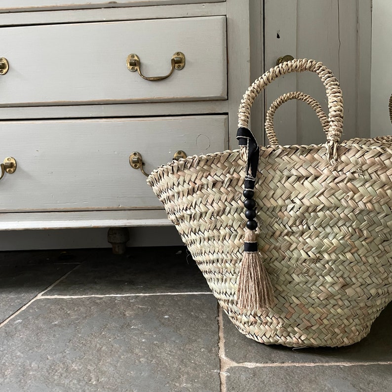 May include: A large, woven straw tote bag with black and white tassel accents. The bag is sitting on a tiled floor in front of a white dresser.