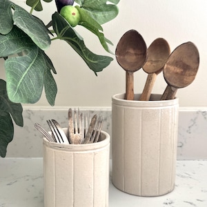 May include: Two off-white ceramic utensil pots with vertical grooves. One contains forks, the other wooden spoons. A fig plant with green leaves and figs is in the background. The utensils are vintage and show signs of wear.