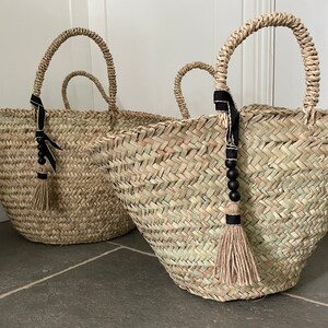 May include: Two large woven straw baskets with black ribbon handles and black bead tassels. The baskets are sitting on a gray tiled floor.
