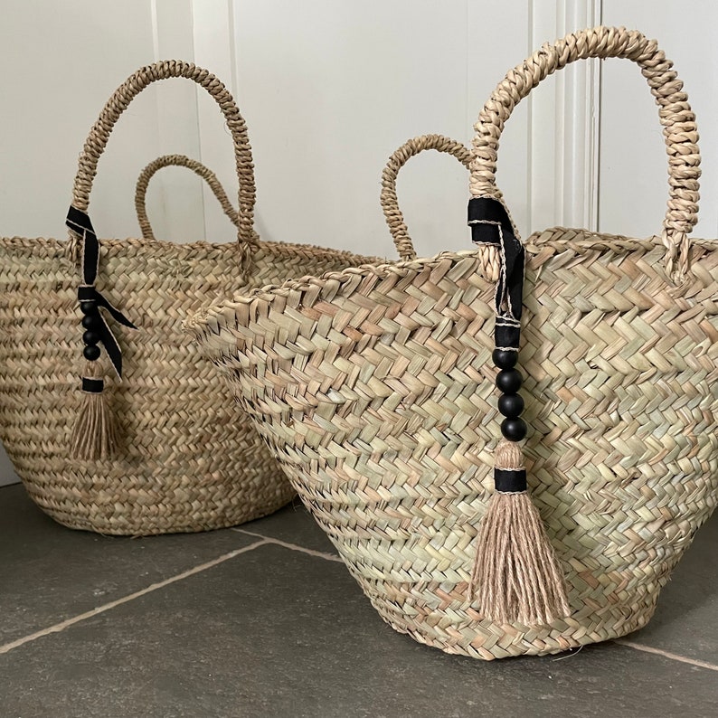 May include: Two large woven straw baskets with black ribbon handles and black bead tassels. The baskets are sitting on a gray tiled floor.