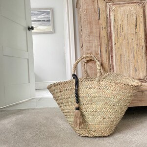 May include: A large, natural woven basket with a black tassel and beads. The basket is sitting on a carpeted floor in front of a wooden cabinet.