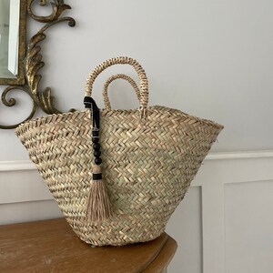 May include: A large, natural woven straw tote bag with two handles. The bag has a black tassel with wooden beads hanging from one of the handles.