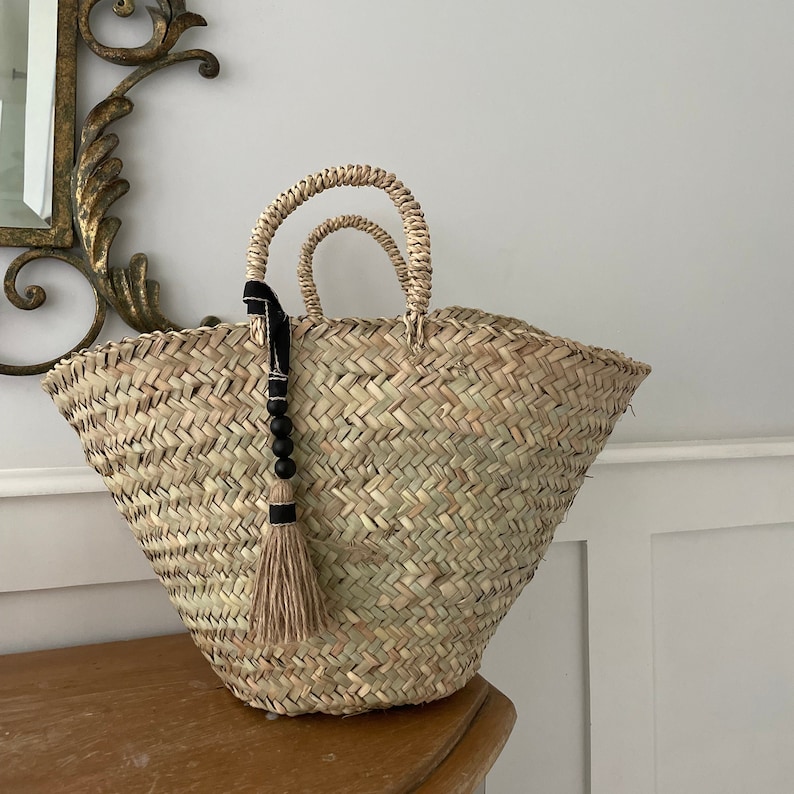 May include: A large, natural woven straw tote bag with two handles. The bag has a black tassel with wooden beads hanging from one of the handles.