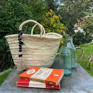 May include: A woven straw tote bag with a black tassel and beaded strap. The bag is sitting on a table with two books and a glass bottle.