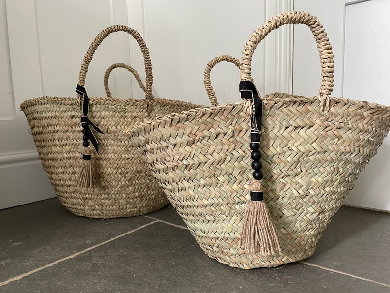 May include: Two large woven straw tote bags with black ribbon handles and black bead tassels. The bags are sitting on a gray tiled floor.