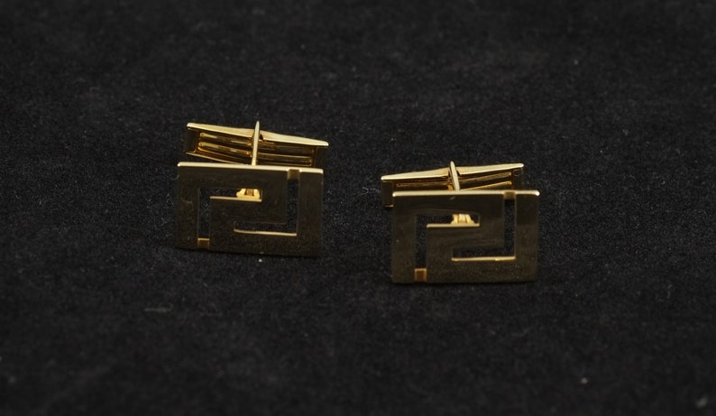 May include: A pair of gold cufflinks with a geometric design. The cufflinks are square shaped and have a Greek key pattern.