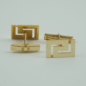 May include: A pair of gold cufflinks with a geometric design. The cufflinks are made of a shiny metal and have a square shape.