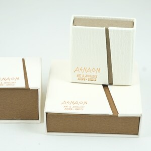 May include: Three white boxes with brown lids. The boxes are stacked on top of each other. The top box has the text "AENAON ART & JEWELLERY ATHENS GREECE" printed on it. The middle box has the text "AENAON ART & JEWELLERY ATHENS GREECE" printed on it. The bottom box has the text "AENAON ART & JEWELLERY ATHENS GREECE" printed on it.