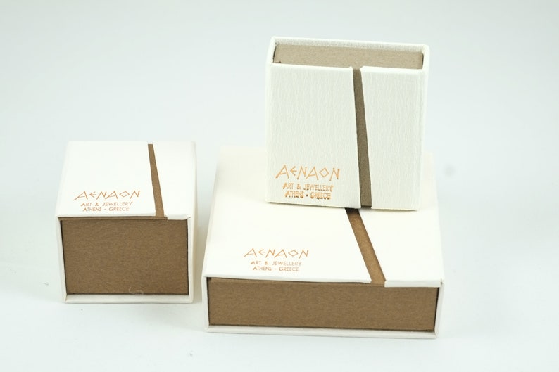 May include: Three white boxes with brown lids. The boxes are stacked on top of each other. The top box has the text "AENAON ART & JEWELLERY ATHENS GREECE" printed on it. The middle box has the text "AENAON ART & JEWELLERY ATHENS GREECE" printed on it. The bottom box has the text "AENAON ART & JEWELLERY ATHENS GREECE" printed on it.