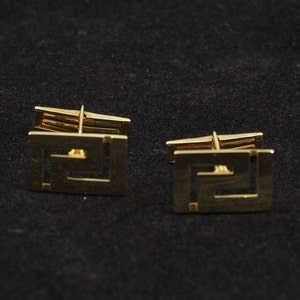 May include: A pair of gold cufflinks with a geometric design. The cufflinks are square shaped and have a detailed pattern.