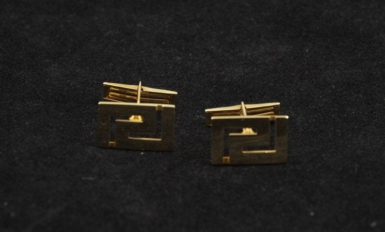 May include: A pair of gold cufflinks with a geometric design. The cufflinks are square shaped and have a detailed pattern.