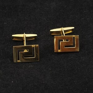 May include: A pair of gold cufflinks with a geometric design. The cufflinks are square shaped with a Greek key pattern.