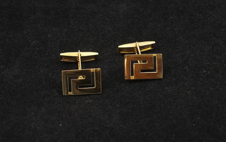 May include: A pair of gold cufflinks with a geometric design. The cufflinks are square shaped with a Greek key pattern.