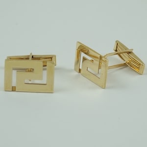 May include: A pair of gold cufflinks with a geometric design. The cufflinks are made of a shiny metal and have a square shape.