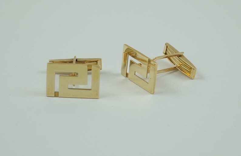 May include: A pair of gold cufflinks with a geometric design. The cufflinks are made of a shiny metal and have a square shape.