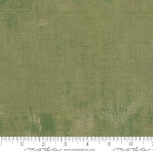 May include: A close-up of a fabric with a distressed, textured appearance. The fabric has a mottled, olive green color with lighter, beige-toned areas. Horizontal and vertical lines create a subtle grid pattern. A ruler is at the bottom.