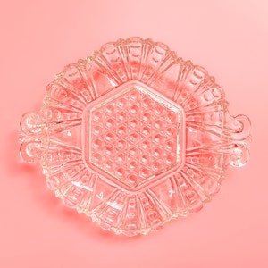 Puede incluir: Un plato de vidrio transparente con un centro hexagonal y un borde decorativo festoneado. El plato está sobre un fondo rosa.