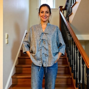 May include: A light blue, textured, loose-fitting blouse with three-quarter length sleeves and a mandarin collar. The front features a vertical placket with four sets of thin, matching fabric ties that are tied in bows, paired with blue denim jeans.
