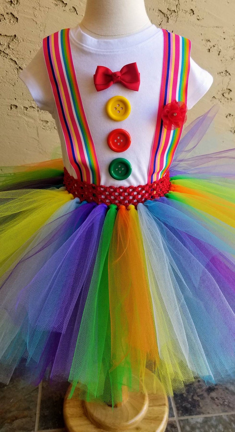 Puede incluir: Un tut&uacute; arco&iacute;ris con una camisa blanca con una pajarita roja y botones de colores. La camisa tiene tirantes arco&iacute;ris.