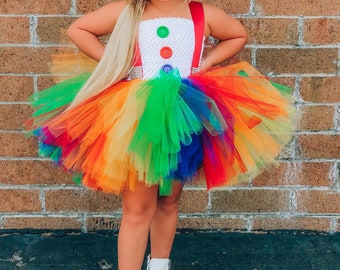 Puede incluir: Una niña pequeña con un disfraz de tutú arcoíris con un top blanco y tirantes rojos. El top tiene tres botones grandes y un diseño de crochet blanco. La niña lleva zapatos blancos y tiene el pelo largo y rubio.