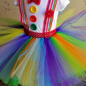 Puede incluir: Un tut&uacute; arco&iacute;ris con una camisa blanca con una pajarita roja, tres botones y tirantes arco&iacute;ris. El tut&uacute; est&aacute; hecho de tul y tiene una cintura roja de ganchillo.