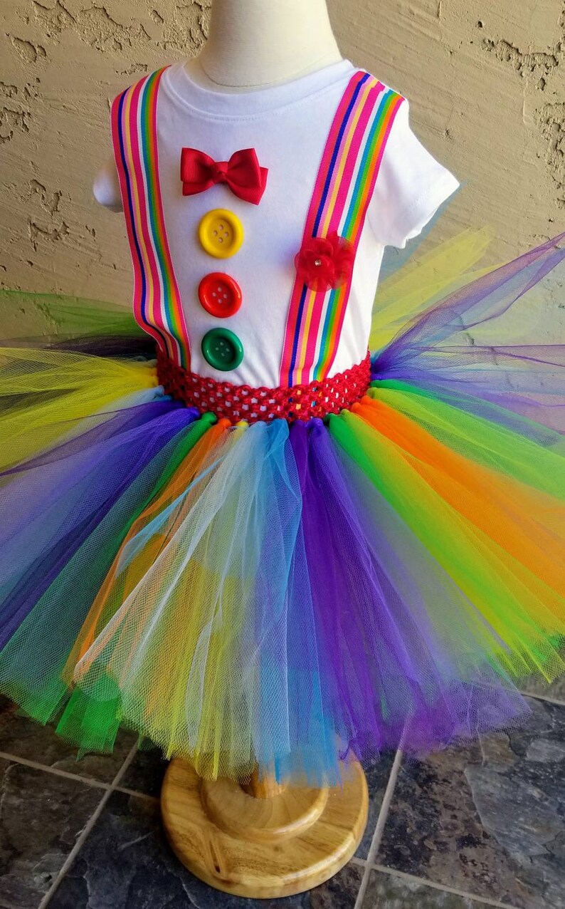 Puede incluir: Un tut&uacute; arco&iacute;ris con una camisa blanca con una pajarita roja, tres botones y tirantes arco&iacute;ris. El tut&uacute; est&aacute; hecho de tul y tiene una cintura roja de ganchillo.