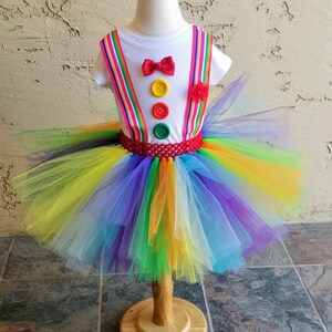 Puede incluir: Un tut&uacute; arco&iacute;ris con una camisa blanca con una pajarita roja y botones de colores. La camisa tiene tirantes rojos y blancos a rayas.