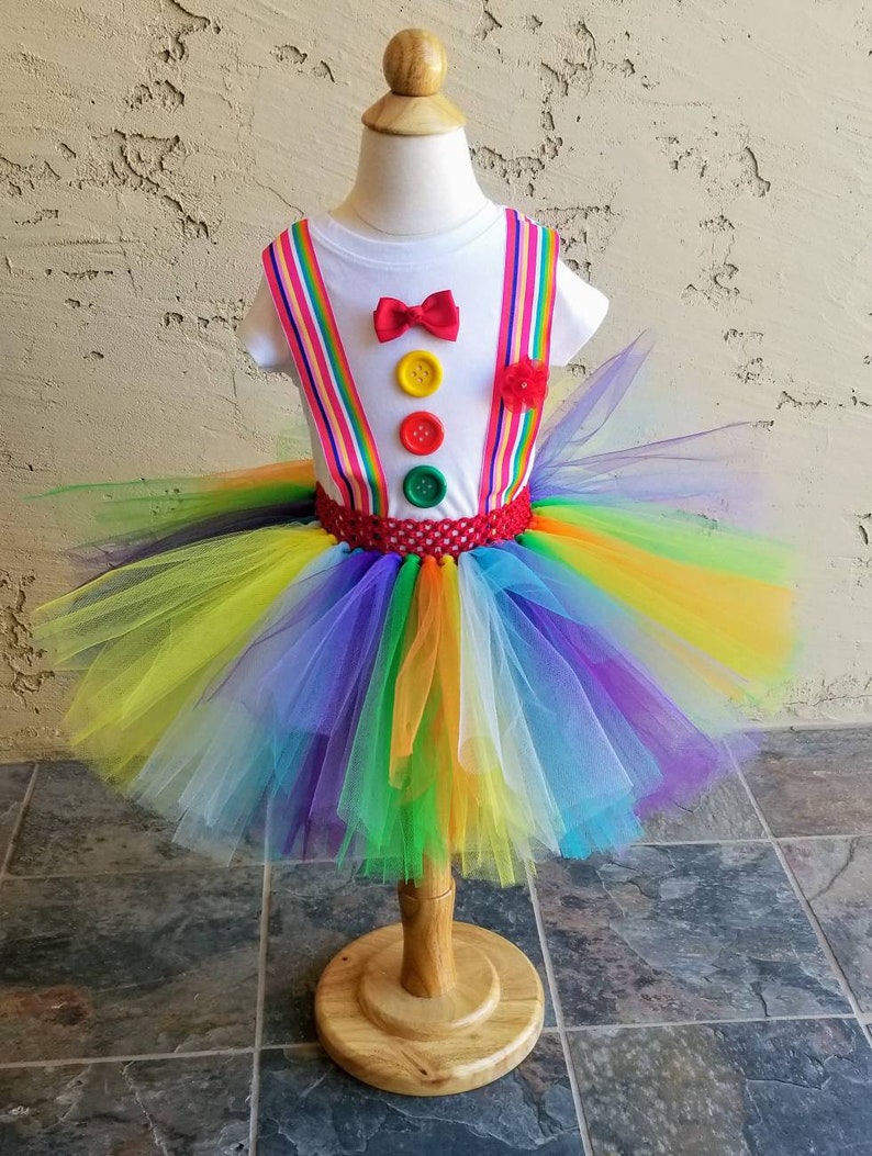 Puede incluir: Un tut&uacute; arco&iacute;ris con una camisa blanca con una pajarita roja y botones de colores. La camisa tiene tirantes rojos y blancos a rayas.