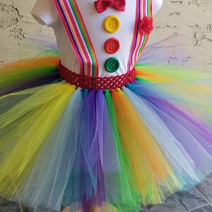 Puede incluir: Un tut&uacute; arco&iacute;ris con una camisa blanca con una pajarita roja y tres botones de colores. La camisa tiene tirantes rojos y de rayas arco&iacute;ris.