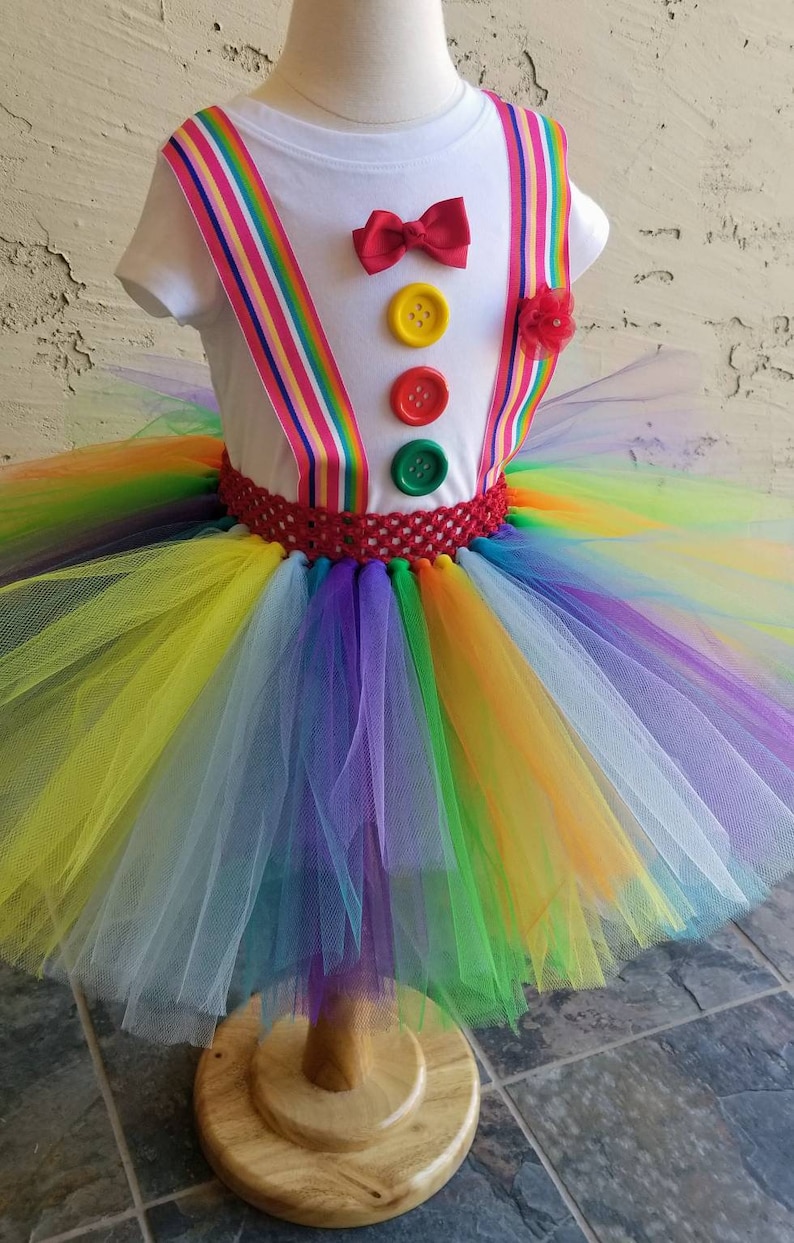 Puede incluir: Un tut&uacute; arco&iacute;ris con una camisa blanca con una pajarita roja y tres botones de colores. La camisa tiene tirantes rojos y de rayas arco&iacute;ris.