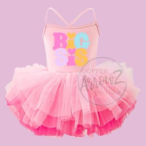 May include: A pink tutu dress with spaghetti straps and a layered skirt. The bodice features the text "BIG SIS" in pastel colors. The dress is designed for a child.