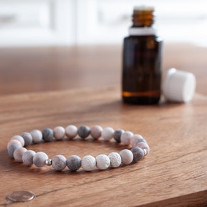 May include: A bracelet made of pink, grey, and white stone beads. The bracelet is displayed on a wooden surface, with a small silver bead and a brown essential oil bottle in the background.