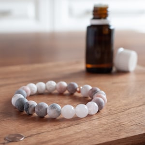 May include: A bracelet made of pink, grey, and white beads with a rose quartz bead in the centre. The bracelet is on a wooden surface.