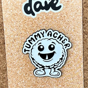 May include: A white enamel pin featuring a cartoon character with a round body, large eyes, and a single tooth, surrounded by the text "TUMMY ACHER". The pin is attached to a rectangular display card with "Pins by dave" and "@thatfelladave" printed on it.
