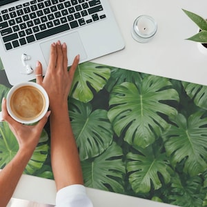 May include: A white ceramic coffee mug with a brown coffee drink inside, sitting on a green and black tropical leaf patterned desk mat. A laptop computer and a small potted plant are visible in the background.