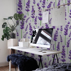 May include: A white wall with a lavender floral wallpaper pattern. A desk with a computer monitor, a keyboard, and a mouse is in front of the wall. A black furry stool is in front of the desk. A bed with a grey duvet is in the foreground.
