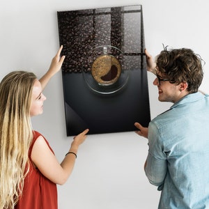Könnte beinhalten: Ein Paar hängt einen gerahmten Druck eines Kaffebechers an einer weißen Wand. Der Druck zeigt eine Nahaufnahme eines Kaffebechers, der mit Kaffee gefüllt ist und von Kaffeebohnen umgeben ist.