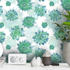 May include: Watercolor-style wallpaper featuring a repeating pattern of green and blue succulent plants on a white background. A white vase, a small radio, and a potted plant with large green leaves sit on a white surface.