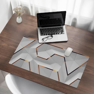 Op de afbeelding: Een grijze en witte geometrische bureauonderlegger met gouden accenten. De onderlegger ligt op een houten bureau met een laptop, een bril en een muis.