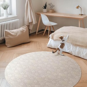 Può includere: Un tappeto rotondo color beige chiaro con un delicato motivo floreale. Un gatto cammina sul tappeto in una stanza con una scrivania in legno, una sedia e un letto.