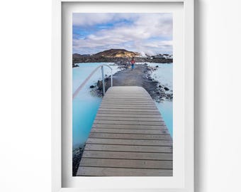 May include: A wooden walkway leads to a blue lagoon in a scenic landscape. The walkway is made of light brown wood planks and has a metal railing on one side. The lagoon is a bright blue colour and is surrounded by rocks and vegetation.
