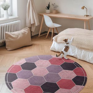 May include: A pink and purple geometric patterned rug with a hexagonal design. The rug is on a hardwood floor with a cat walking on it.