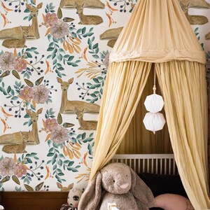 Puede incluir: Una habitación de bebé blanca con una cama con dosel que presenta un dosel beige y una cuna blanca. La pared está cubierta con un papel tapiz con un patrón repetitivo de ciervos de acuarela y diseños florales en tonos de rosa, naranja y verde.