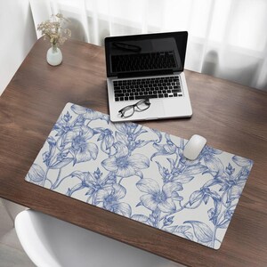 Puede incluir: Una alfombrilla de escritorio blanca con un patrón floral azul. La alfombrilla está sobre un escritorio de madera con un portátil, gafas y un ratón blanco.