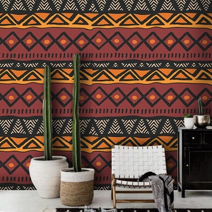 May include: A living room interior with a patterned wallpaper featuring geometric shapes in orange, red, and black. Two tall potted plants stand in front of the wall, and a white chair with a black throw blanket sits in front of the plants. A black rug with a geometric pattern covers the floor.