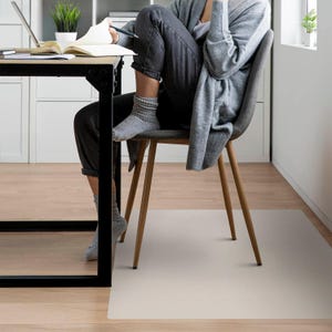May include: A light beige chair mat placed on a wooden floor. A person is sitting in a wooden chair with a grey cardigan and black trousers. The chair is positioned over the mat.
