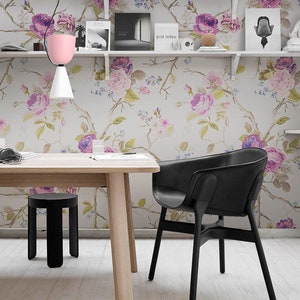 May include: A black leather chair with a wooden frame sits at a wooden table with a black stool. The table and chair are in front of a wall with a floral wallpaper featuring pink and purple roses. The wall also has white shelves with books and decorative items.