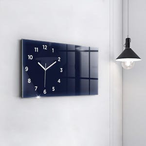 Puede incluir: Un reloj de pared rectangular con una esfera azul oscuro y números blancos. Las manecillas del reloj son blancas. El reloj está montado en una pared blanca. Una lámpara colgante negra cuelga cerca.