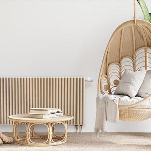 Op de afbeelding: Een witte muur met een beige radiator, een hangstoel van wicker met witte kussens, een salontafel van wicker met boeken, een plant in een pot en een witte kat op een tapijt.